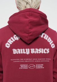 Sweatshirt à capuche bordeaux avec un texte graphique blanc au dos, y compris "DAILY BASICS" et un détail signature. Texture de tissu douce.