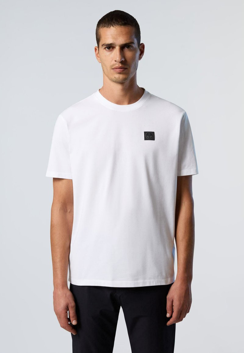 T-shirt bianco in cotone con maniche corte, scollo tondo e un piccolo logo nero applicato sul petto. Texture liscia e design minimalista.