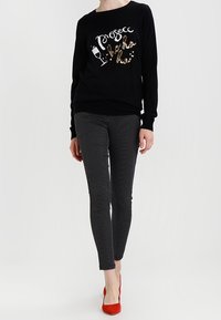 Schwarzer Pullover mit glitzerndem "Prosecco"-Text, kombiniert mit taillierten schwarz-weißen gepunkteten Hosen. Helle rote High Heels runden das Outfit ab.