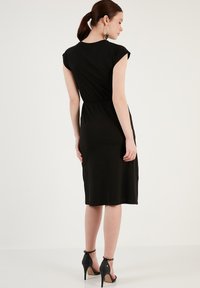 Robe noire, longueur genou avec une taille ajustée et des manches courtes, fabriquée dans un tissu lisse. Portée avec des sandales à talons hauts noires.