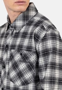 Zwart-wit geruit overhemd met een knoopsluiting, grote kraag en een voorzak met logo. Zachte stof met een gestructureerde afwerking.