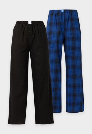 Femme debout pieds nus portant un haut noir à manches longues et un pantalon large à carreaux bleus, les mains dans les poches, sur fond blanc.