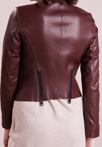 Bordeauxfarbene Lederjacke mit taillierter Passform, ausgestattet mit zwei sichtbaren Reißverschlüssen auf der Rückseite und einem kontrastierenden cremefarbenen Stoff am unteren Rand.