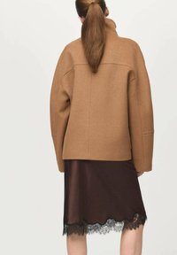 Chaqueta grande de mezcla de lana marrón con cuello alto, combinada con una falda de satén marrón con un borde de encaje negro, vista desde atrás.