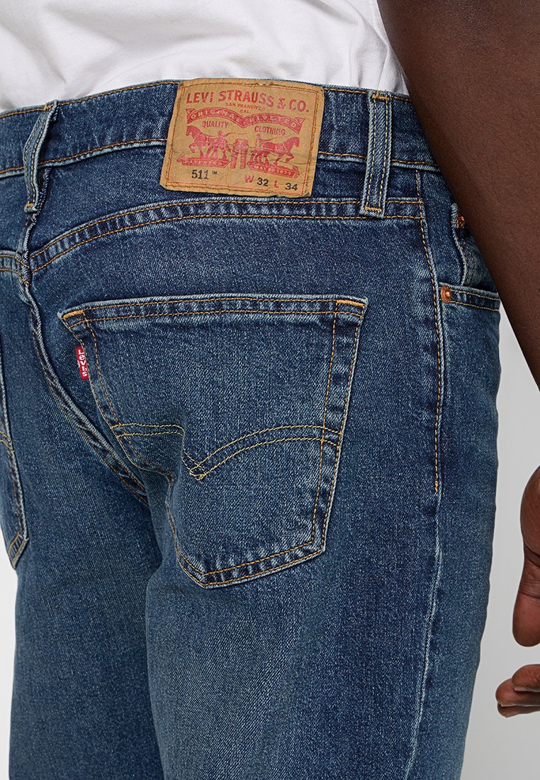 Jean en denim bleu foncé avec un léger délavé, présentant un patch en cuir marron à la taille avec des lettres rouges. Design à cinq poches avec une couture classique.