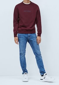 Felpa color borgogna con scollo a girocollo, polsini e orlo a costine; presenta il logo "PEPE JEANS" in stampa scura. Abbinata a jeans azzurri e sneaker bianche.