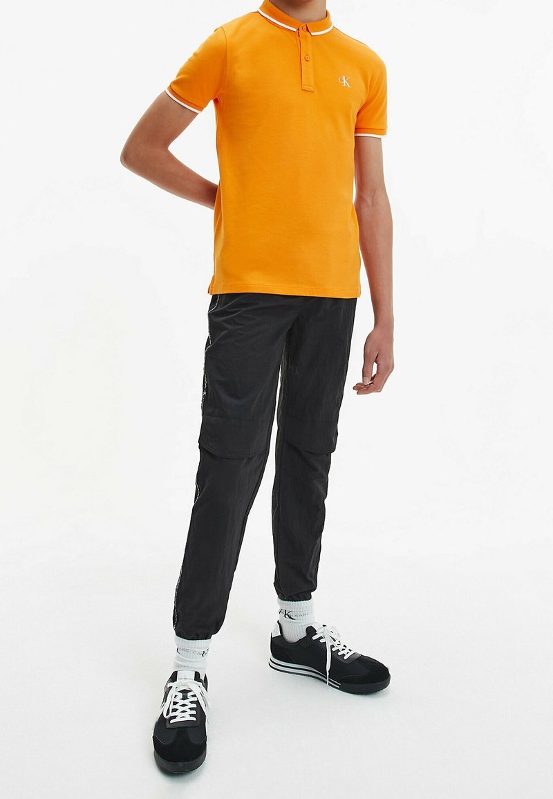 Oranžové polo tričko s bílými pruhy na límci, spárované s černými cargo kalhotami a černými teniskami s bílými tkaničkami a akcenty.