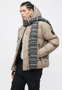 Μπεζ μπουφάν με φουσκωτό ύφασμα, ψηλό γιακά, τσέπες με φερμουάρ και μαύρο λογότυπο στο μανίκι· συνδυασμένο με ριγέ γκρι κασκόλ.