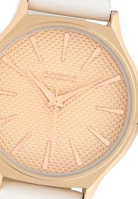 Gold-tone watch with a textured peach dial, raised hour markers, narrow hands, and a white silicone strap. Circular case design.