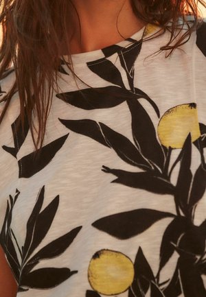 Biała koszulka z tkaniny z czarnymi wzorami liści i żółtymi akcentami cytryn. Koszulka ma luźny krój i okrągły dekolt.