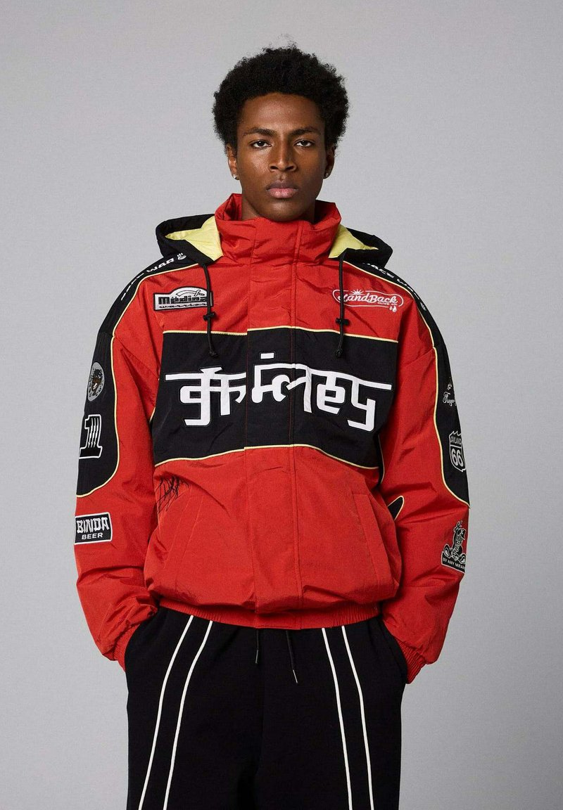 Giacca puffer rossa con accenti neri e foderatura gialla. Presenta toppe e dettagli testuali sulla parte anteriore e sulle maniche. Cappuccio con coulisse.