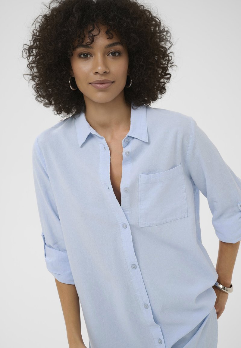 Gaiši zila pogājama krekla ar mīkstu audumu, kas ir ar krūšu kabatu un uzrullētām piedurknēm, taisna griezuma ar smalku faktūru.