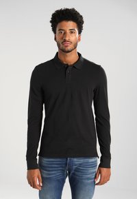 Zwarte polosweater met lange mouwen, een platte kraag en een doorlopende knoopsluiting met drie knopen, gecombineerd met blauwe spijkerbroek. De stof oogt glad en aansluitend.