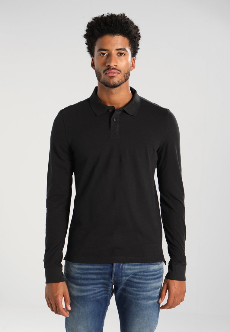Zwarte polosweater met lange mouwen, een platte kraag en een doorlopende knoopsluiting met drie knopen, gecombineerd met blauwe spijkerbroek. De stof oogt glad en aansluitend.
