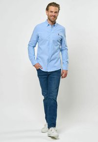 Világoskék ing, gallérral, sötétkék farmernadrággal párosítva. Az ing elején egy kis embléma található, hosszú ujjú.