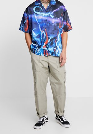 Personne portant une chemise à manches courtes bleu et rouge à motif de flammes, un pantalon cargo beige et des baskets noires et blanches, debout les mains dans les poches.