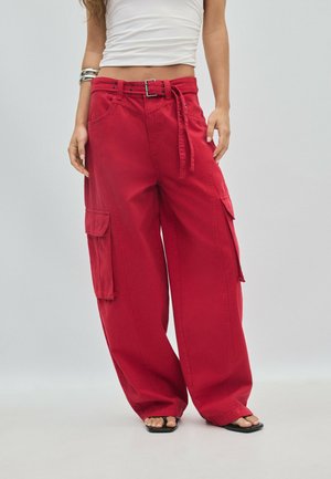 Femme portant un pantalon cargo rouge à jambes larges avec une ceinture, un top court blanc, un bracelet en argent et des sandales noires à bout ouvert.