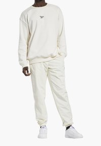 Hombre vestido con un sudadera off-white de Reebok, pantalones a juego y zapatillas blancas con calcetines negros, de pie con una mano en el bolsillo.