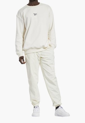 Uomo che indossa un felpa beige chiaro Reebok, pantaloni abbinati e sneakers bianche con calzini neri, in piedi con una mano in tasca.