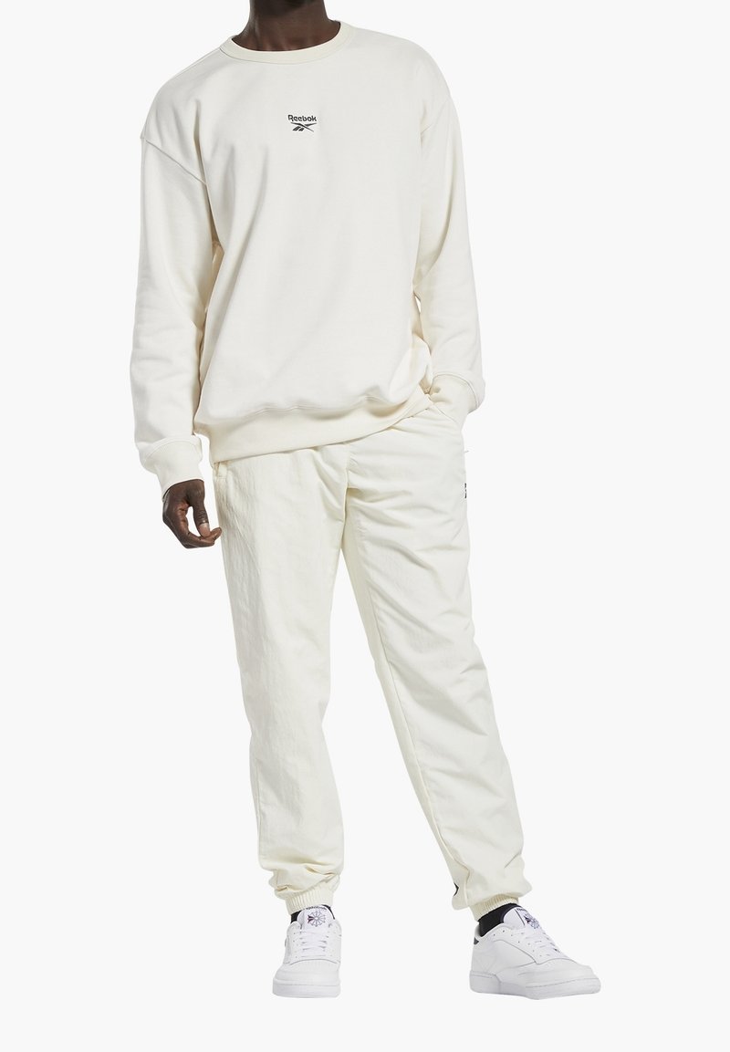 Hombre vestido con un sudadera off-white de Reebok, pantalones a juego y zapatillas blancas con calcetines negros, de pie con una mano en el bolsillo.