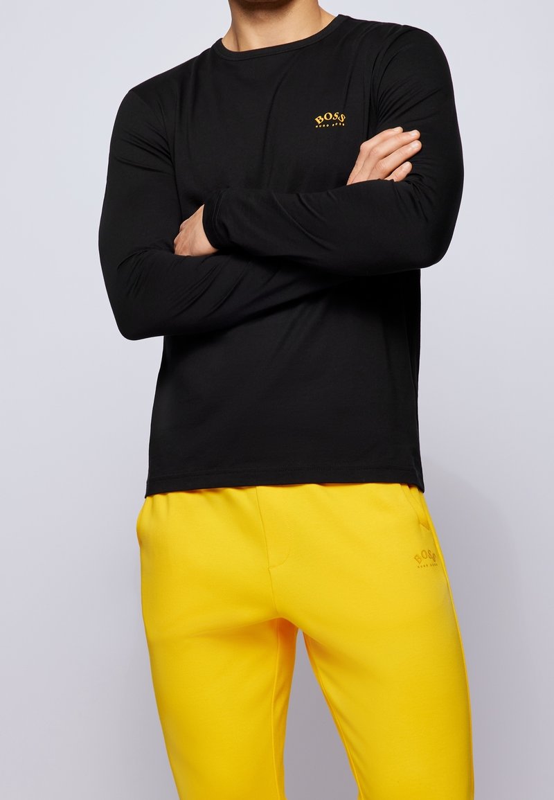 Schwarzes Langarmshirt mit rundem Ausschnitt und kleinem gelben Logo, kombiniert mit leuchtend gelben Jogginghosen, die ein passendes Logo tragen.