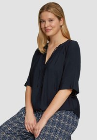 Marineblauwe blouse met korte mouwen, een V-hals en trekkoorddetails. Gladde textuur met een casual pasvorm, gecombineerd met gestreepte blauw en crèmekleurige broeken.