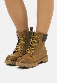 Botas de tobillo de cuero marrón con cordones dorados, parte superior de cuero texturizado, cremallera lateral y suela de goma gruesa con patrón de agarre.