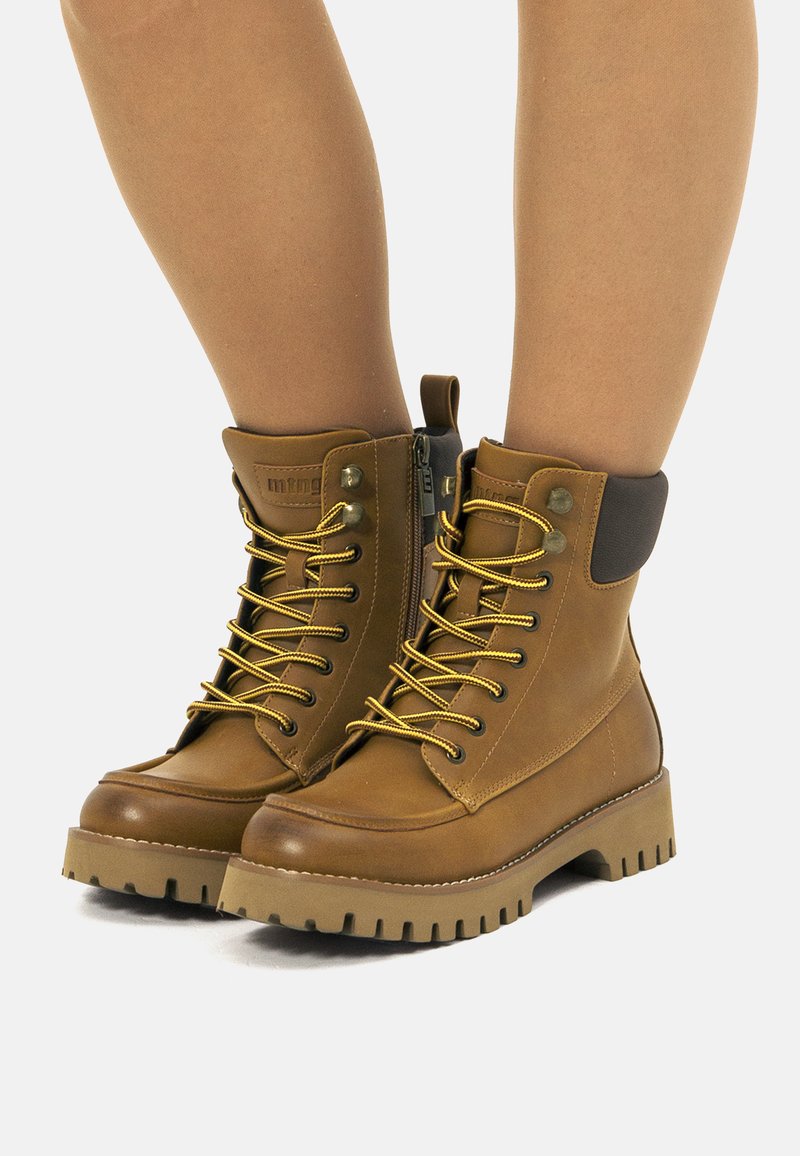 Botas de tobillo de cuero marrón con cordones dorados, parte superior de cuero texturizado, cremallera lateral y suela de goma gruesa con patrón de agarre.