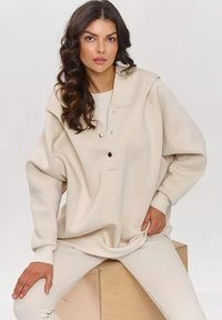 Beżowy, duży sweatshirt z dużym kołnierzem, zapięciem na guziki i ściągaczami na końcach rękawów. Miękka tekstura współgra z dopasowanymi beżowymi legginsami z ściągaczami.