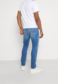 Man i vit T-shirt, ljusblå jeans och vita Reebok-sneakers som står mot vit bakgrund, vänd bort från kameran.