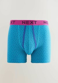 Boxers bleus avec un motif ondulé, une taille rose avec "NEXT" en noir, et des détails de surpiqûre violette. Texture de tissu douce.