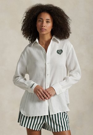 Kvinde med krøllet hår iført en hvid skjorte med knapper med et lille grønt våbenskjold og stribede shorts, stående mod en ensfarvet baggrund.