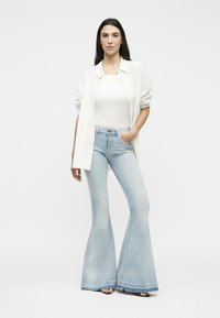 Γυναίκα στέκεται με το ένα χέρι στην τσέπη, φοράει λευκό πουκάμισο, λευκό φανελάκι, ανοιχτό μπλε καμπάνα τζιν και μαύρα πέδιλα με ανοιχτές μύτες σε λευκό φόντο.