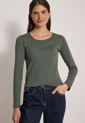 Femme aux longs cheveux foncés portant une chemise à manches longues vert olive et un jean bleu foncé, une main dans la poche, sur un fond uni.