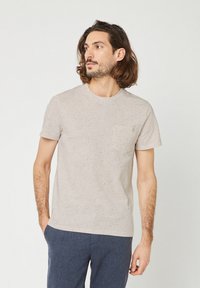 Uomo con capelli alla spalla che indossa una camicia a maniche corte beige maculata con tasca e pantaloni blu, che guarda a sinistra.