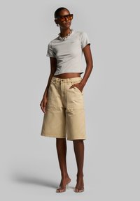 T-shirt grigio chiaro cropped, pantaloni corti di denim beige con grandi tasche, e sandali con tacco trasparente. Gli accessori includono una collana con catena spessa.