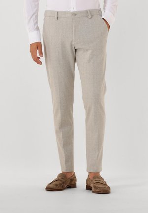 Mann in beigen maßgeschneiderten Hosen mit Bügelfalte, kombiniert mit braunen Wildleder-Mokassins und einem weißen Hemd vor einem schlichten Hintergrund.