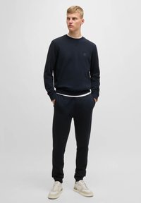 Granatowy sweter z okrągłym dekoltem i ściągaczami na rękawach, w połączeniu z pasującymi dresami. Zauważalne szczegóły obejmują małe logo i białą podszewkę.
