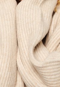 Béžový žebrovaný šál vyrobený z měkkého, strukturovaného materiálu, s podrolovanými okraji a pohodlným, volně splývajícím vzhledem.