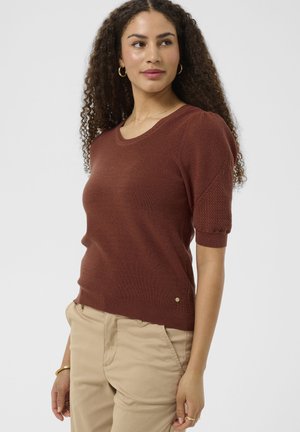 Kvinde med krøllet hår iført en rustfarvet striktop med korte ærmer og beige bukser, ser lidt til siden.