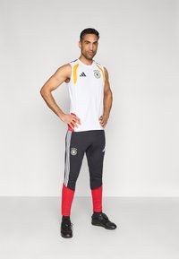 Hvid ærmeløs sportstop med sorte og gule detaljer, kombineret med sorte og røde tætsiddende bukser med hvide striber og logo. Sorte sko.