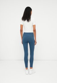 Leggings azuis com uma cintura alta, tecido suave e pernas ajustadas, combinados com uma blusa branca de mangas curtas e ténis brancos.