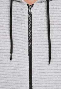 Ljusgrå zip-up hoodie med en strukturerad tyg, svart dragkedja och snörning. Framöppningen har en slät, rak dragkedjelinje.