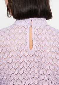 Πουκάμισο από δαντέλα λεβάντας με ψηλό λαιμόκοψη και λεπτομέρεια με άνοιγμα στην πλάτη, περίπλοκα γεωμετρικά σχέδια και υφασμάτινη υφή.