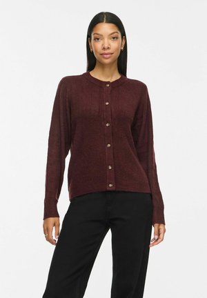 Femme aux longs cheveux raides portant un cardigan marron foncé boutonné et un pantalon noir, debout devant un fond clair uni.