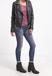 Czarna skórzana kurtka motocyklowa noszona na bordowym t-shircie z grafiką, w zestawieniu z przetartymi niebieskimi dopasowanymi dżinsami i czarnymi botkami na obcasie.