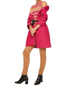 Manteau en laine fuchsia porté sur un haut tricoté à rayures beiges et roses, associé à un pantalon ajusté beige et des bottines noires avec un accent doré.