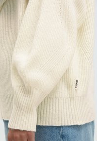 Cremefarbenes, gestricktes Sweatshirt mit geripptem Muster, ausgestattet mit einem Etikett mit der Aufschrift "SENSES" und leicht übergroßen Ärmeln in der Nähe eines Denim-Taillenbunds.