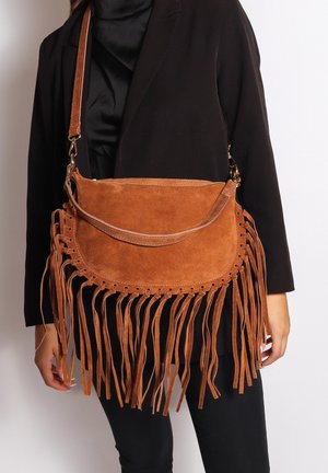 Sac en daim marron porté en bandoulière avec de longs franges, associé à un manteau noir et un pantalon, sur une personne devant un fond uni.