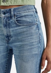 Primer plano de una persona que lleva jeans de mezclilla azul claro y un top verde corto, mostrando detalles de los bolsillos y las costuras.